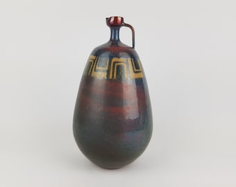May include: A handcrafted ceramic jug with a unique shape and a handle. The jug features a dark red and blue glaze with gold geometric patterns. The jug's design is both functional and decorative, suitable for display or use.