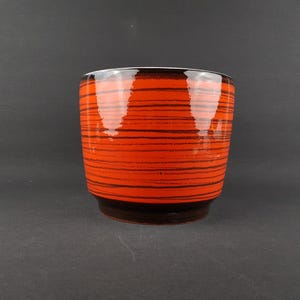 May include: A ceramic planter with a vibrant orange glaze and horizontal black stripes. The planter has a dark brown base and a white interior rim. Suitable for indoor or outdoor use, this piece is a decorative item.