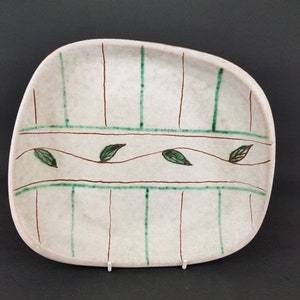 May include: A white ceramic serving dish with a unique, free-form shape. The dish features a decorative design of green leaves and brown and green lines.
