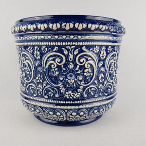 Könnte beinhalten: Blau-weißer Keramik-Blumentopf mit aufwendigen geprägten Blumen- und Rankenmustern. Der Topf hat eine abgerundete Form mit detailliertem Rand und Boden. Das Farbschema ist ein tiefes Blau mit weißen Akzenten.