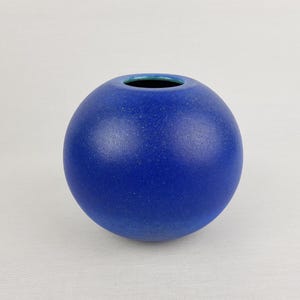 May include: A spherical ceramic vase with a vibrant cobalt blue matte finish and a speckled texture. The vase features a small, circular opening at the top with a smooth, contrasting teal-colored interior rim, creating a simple, rounded form.