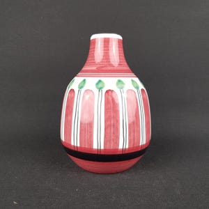 Puede incluir: Un jarrón decorativo de cerámica con una combinación de colores rojo, blanco y negro. El jarrón presenta rayas verticales rojas alternadas con rayas blancas. Formas verdes en forma de hojas están cerca de la parte superior. El jarrón tiene un cuerpo redondeado y un cuello estrecho.