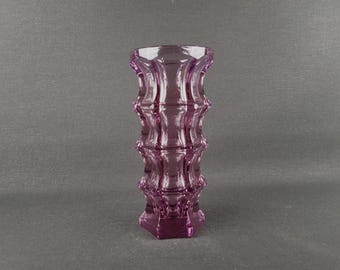 Può includere: Un vaso alto di vetro color ametista con un design unico e strutturato. Il vaso ha una base esagonale e una serie di segmenti arrotondati impilati. Il vetro è trasparente e la forma generale è cilindrica.