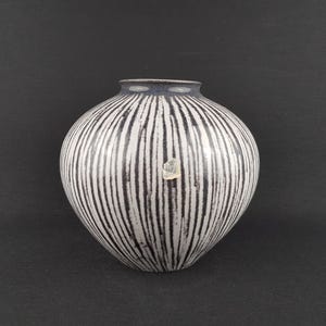 May include: A ceramic vase with a rounded body and a narrow neck. The vase is white with vertical black stripes. The rim is dark blue. A small, clear sticker is affixed to the front of the vase.
