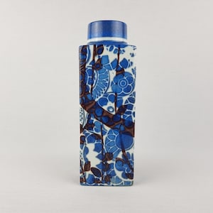 May include: A rectangular ceramic vase with a blue and white floral pattern. The vase has a blue top and a repeating design of stylized flowers and leaves in shades of blue and brown. The vase is a decorative item.