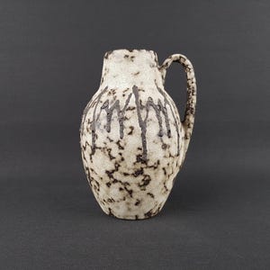 Op de afbeelding: Een keramische kan met een gespikkelde, gebroken witte basis en een donkerbruin druppelpatroon. De kan heeft een handvat en een afgeronde body, geschikt voor het serveren van dranken of als decoratief item. De algehele esthetiek is rustiek en vintage.