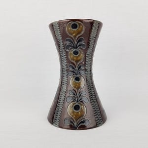 Puede incluir: Un jarrón de cerámica marrón con una forma de reloj de arena única. El jarrón está decorado con un patrón repetitivo de flores y hojas estilizadas en tonos marrones, dorados y grises.