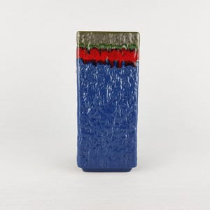 Puede incluir: Un jarrón rectangular de cerámica con cuerpo azul texturizado. La sección superior presenta bandas rojas, verdes y marrones. El jarrón tiene un diseño moderno y geométrico, adecuado para la decoración del hogar.