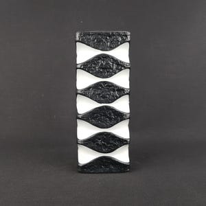 Könnte beinhalten: Eine rechteckige Keramikvase mit kontrastreichem Design, bestehend aus einem glatten weißen Grundkörper und fünf horizontalen, texturierten schwarzen Bändern. Die schwarzen Bänder haben eine raue, organische Oberfläche und eine geschwungene Form.