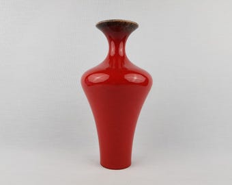 May include: A tall, glossy red vase with a flared base and a narrow neck. The rim of the vase is a dark brown color. The vase has a smooth, reflective surface and is set against a plain white background.