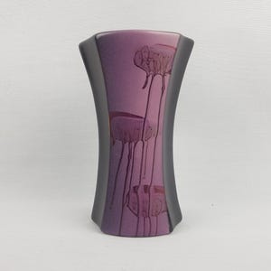 May include: A tall, curved vase with a matte black finish on the sides and a central panel featuring a gradient of purple and pink. The central panel is decorated with a dark, dripping paint design that resembles abstract floral or jellyfish shapes.