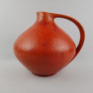 Puede incluir: Jarra de cerámica rojo-naranja hecha a mano, con cuerpo redondeado y asa curva. La jarra tiene un acabado liso y brillante y un pequeño pico. El diseño es sencillo y elegante, adecuado para servir bebidas o como pieza decorativa.