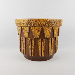 May include: A brown and yellow ceramic planter with a textured surface. The planter has a cylindrical shape with a wide rim and a series of vertical and triangular patterns around the body. The planter is suitable for indoor or outdoor use.