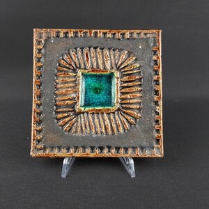 May include: A square ceramic tile featuring a dark charcoal grey textured border with a repeating notched edge. The centre contains a recessed square glazed in vibrant turquoise blue, surrounded by a radiating pattern of raised brown ridges.