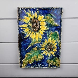 May include: A rectangular ceramic wall plaque featuring a relief design of two yellow sunflowers with brown centers, surrounded by green leaves against a deep blue, textured background with a mottled finish and a rough, uneven border.