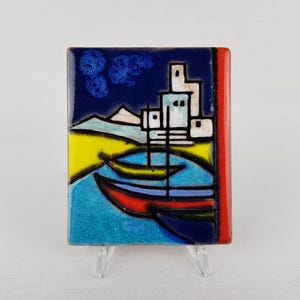 Op de afbeelding: Een rechthoekige keramische tegel met een kustscène. Het kunstwerk toont boten op turquoise water, een gele kustlijn en gebouwen onder een donkerblauwe lucht met gestileerde wolken. De tegel heeft een rode rand.
