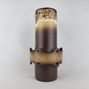 Könnte beinhalten: Eine zylindrische Keramikvase mit dunkelbrauner matter Oberfläche. Der obere Rand zeigt eine gesprenkelte cremefarbene und braune Glasur, während ein zentrales Zierband mit gewelltem Rand und kreisförmigen Aussparungen eine gesprenkelte beige-braune Optik aufweist.