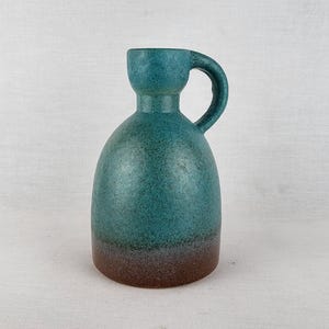 May include: A ceramic pitcher with a rounded body and a single handle. The upper portion features a speckled teal glaze, while the base transitions into a solid brown tone. The vessel has a narrow neck and a wide, bulbous bottom section.