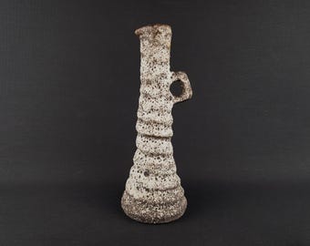 May include: A tall, textured ceramic vase with a handle. The vase has a light beige and brown speckled pattern, with a flared base and a narrow neck. The handle is a small loop. The vase is approximately 12 inches tall.