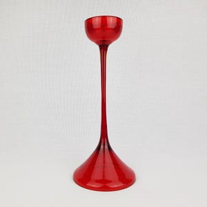 Op de afbeelding: Een hoge, rode glazen kaarsenstandaard met een brede, uitlopende basis en een smalle, cilindrische bovenkant.