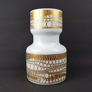 May include: White ceramic vase with a gold circular and dot pattern. The vase has a wide base, a narrow neck, and a flared top. The gold pattern is repeated around the top and bottom, creating a decorative design.