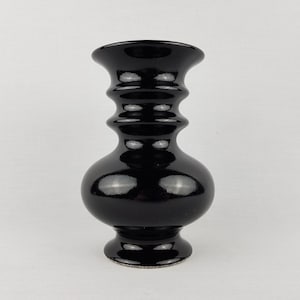 Puede incluir: Un jarrón negro brillante con una forma distintiva. Presenta una base redondeada, una serie de ranuras horizontales y un borde acampanado. El jarrón es probablemente de cerámica o vidrio y tiene una superficie reflectante. El diseño es elegante y sencillo.