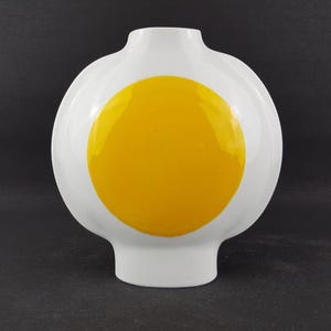 Op de afbeelding: Een witte keramische vaas met een bolvormig lichaam en een smalle hals. De voorzijde van de vaas is voorzien van een groot, effen, felgeel cirkelvormig ontwerp dat in het midden van het oppervlak is geplaatst. De vaas heeft een gladde afwerking.