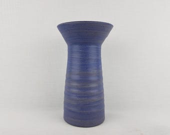 May include: A blue ceramic vase with a flared top and a cylindrical body. The vase has a textured surface with horizontal ridges and a speckled appearance. The colour is a deep, muted blue, suitable for home decor.