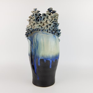 May include: A tall ceramic vase featuring a dark black base that transitions into a bright blue drip glaze over a cream-colored upper section. The top of the vase is adorned with a dense cluster of small, sculpted, mushroom-like ceramic forms.
