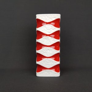 May include: A white ceramic vase with a red and white geometric pattern. The vase has a textured surface and a rectangular shape.