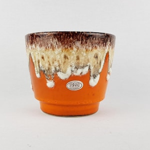 Puede incluir: Una maceta de cerámica naranja con un diseño en capas. El borde superior es marrón oscuro, que pasa a gotas beige y blancas sobre la base naranja. Una pequeña pegatina ovalada con texto es visible en la parte delantera.