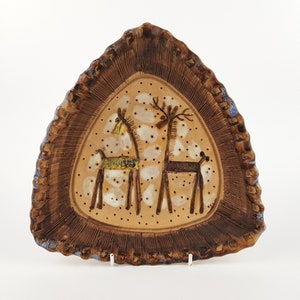 May include: A triangular ceramic plate with a textured dark brown rim and a light tan centre. The centre features a hand-painted illustration of two stylised deer figures surrounded by small white and brown dots on a smooth, glazed surface.