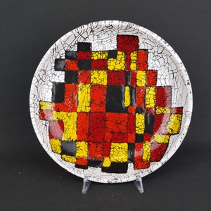 May include: A white ceramic plate with a crackle glaze finish. The plate is decorated with a geometric pattern of red, yellow, and black squares.