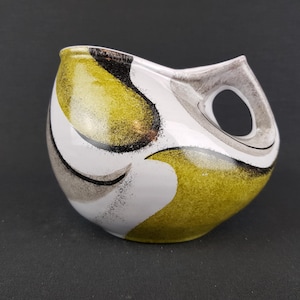 May include: A white ceramic pitcher with a unique abstract design featuring black, gray, and yellow accents. The pitcher has a wide, rounded body and a handle with a circular opening.