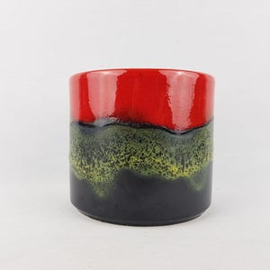 Może przedstawiać: Cylindryczny ceramiczny wazon z błyszczącym wykończeniem. Góra jest czerwona, przechodząca w pas zieleni i żółci, a następnie w czarną podstawę. Wazon jest prawdopodobnie przeznaczony do celów dekoracyjnych.