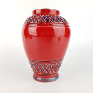 May include: A red ceramic vase with blue and white geometric designs around the top and bottom. The vase has a rounded body and a wide opening.