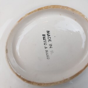 May include: A white ceramic plate with a slightly irregular, light brown rim. The words "MADE IN IT. FATTO A MANO" are printed in black on the center of the plate. The plate has a simple, elegant design.