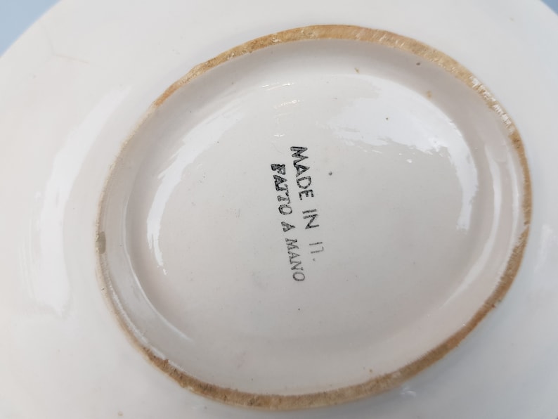 May include: A white ceramic plate with a slightly irregular, light brown rim. The words "MADE IN IT. FATTO A MANO" are printed in black on the center of the plate. The plate has a simple, elegant design.