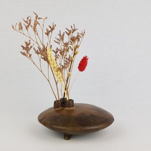 Puede incluir: Un jarrón de madera marrón oscuro con una forma única, con tres aberturas para flores secas. El jarrón está lleno de flores secas, incluyendo una flor roja, una espiga de trigo amarilla y ramas marrones.