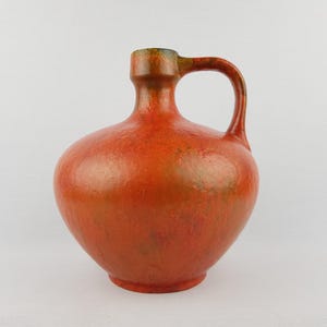 Puede incluir: Una jarra de cerámica redonda de color rojo anaranjado con asa. La jarra tiene un cuerpo bulboso y un cuello estrecho. El asa es curva y está unida al cuello y al cuerpo. La jarra está esmaltada con un acabado moteado.