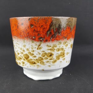 Puede incluir: Una maceta de cerámica con superficie texturizada. La sección superior es de color naranja vibrante, que pasa a blanco con motas marrones y una base blanca. La maceta tiene una forma redondeada única.