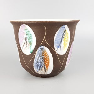 May include: A brown ceramic cup with a white rim and a design of colorful leaves. The cup is decorated with a pattern of gold lines.