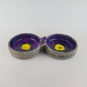 May include: A double-bowl dish featuring a rough, gray, stone-like exterior texture. The interior of each bowl is finished with a smooth, glossy glaze in deep purple, transitioning to a bright yellow center. The two bowls are joined by a small, purple, translucent glass accent piece.