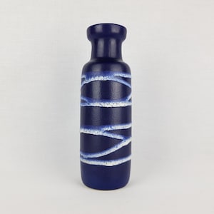 Puede incluir: Un jarrón de cerámica azul oscuro con cuerpo cilíndrico y cuello estrecho. El jarrón presenta bandas horizontales blancas y azul claro, creando un diseño abstracto texturizado. El jarrón mide unos 25 cm de alto.