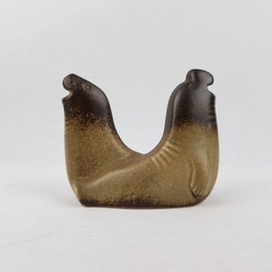 Könnte beinhalten: Eine Keramikskulptur von zwei Robben, die an der Basis verbunden sind und eine U-Form bilden. Die Robben sind hellbraun mit dunkelbraunen Köpfen. Die Skulptur hat eine strukturierte Oberfläche und steht vor einem weißen Hintergrund.