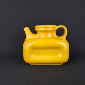 May include: A bright yellow ceramic teapot with a rounded body and a single handle. The teapot has a unique design with a raised rectangular section on the front.