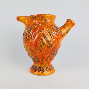 May include: A ceramic pitcher with a textured orange and brown glaze. The pitcher has a spout and a round body with a flared base.