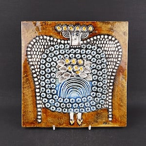 Könnte beinhalten: Quadratische Keramikfliese mit einem stilisierten Engel-Design. Der Engel hat ein weißes Gesicht, eine Krone und große Flügel mit einem Muster aus weißen Punkten und blauen Kreisen. Die Fliese hat einen braunen Rand und einen dunklen Hintergrund.