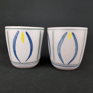 May include: Two ceramic mugs with a white background and blue and yellow abstract designs. The mugs have a green rim and a gray stripe around the bottom.