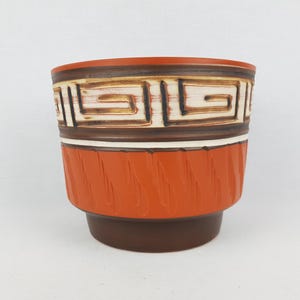 May include: A brown and orange ceramic planter with a geometric design. The planter has a white band with a brown and white geometric pattern around the top.